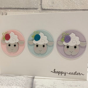 Op de afbeelding: Een witte Paas wenskaart met drie cirkelvormige schapenontwerpen in pastelkleuren. Elk schaap heeft een pluizige witte kop, een grijs gezicht en een bloemendetail. De woorden "happy easter" zijn in cursief geschreven.