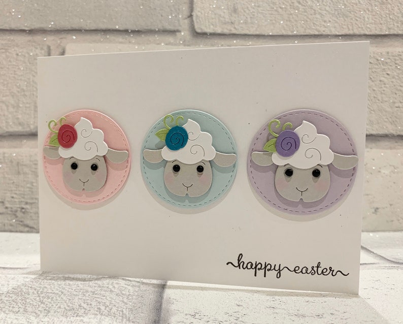 Op de afbeelding: Een witte Paas wenskaart met drie cirkelvormige schapenontwerpen in pastelkleuren. Elk schaap heeft een pluizige witte kop, een grijs gezicht en een bloemendetail. De woorden "happy easter" zijn in cursief geschreven.