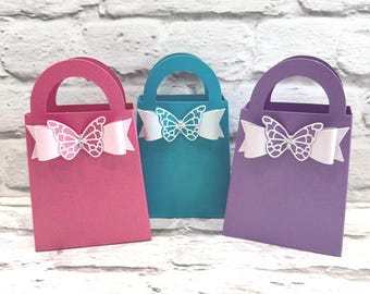 Puede incluir: Tres bolsas de regalo coloridas en rosa, turquesa y morado. Cada bolsa tiene un asa, una cinta blanca y una mariposa decorativa. La mariposa tiene un diseño calado delicado.