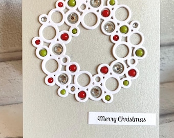 Puede incluir: Una tarjeta de felicitación gris claro con una corona blanca troquelada hecha de círculos interconectados. La corona está decorada con pequeñas cuentas rojas y verde lima y pedrería transparente. Una etiqueta blanca dice Merry Christmas.