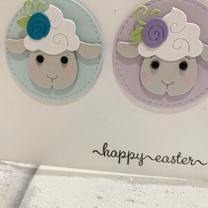 Op de afbeelding: Witte kaart met drie schapenillustraties op pastelkleurige cirkels met gestikte randen. De schapen hebben bloemendetails. De tekst "happy easter" staat in sierlijke letters op de kaart.