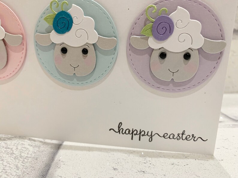 Op de afbeelding: Witte kaart met drie schapenillustraties op pastelkleurige cirkels met gestikte randen. De schapen hebben bloemendetails. De tekst "happy easter" staat in sierlijke letters op de kaart.