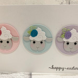 Op de afbeelding: Een witte kaart met drie schapenillustraties in pastel cirkels. Elk schaap heeft een pluizige witte kop, een grijs gezicht en een bloem. De woorden "happy easter" zijn in cursief geschreven.