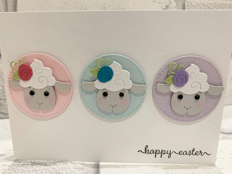 Op de afbeelding: Een witte kaart met drie schapenillustraties in pastel cirkels. Elk schaap heeft een pluizige witte kop, een grijs gezicht en een bloem. De woorden "happy easter" zijn in cursief geschreven.