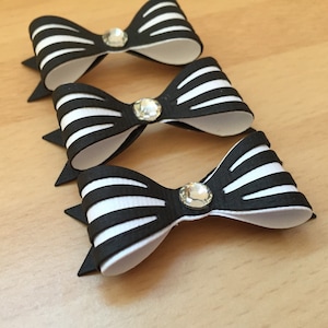 May include: Three black and white striped bow embellishments with a sparkling rhinestone centre. The bows are arranged in a row, with the top bow slightly out of focus. The bows are on a light brown surface.
