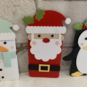 Puede incluir: Tres recortes de papel con tem&aacute;tica navide&ntilde;a: un mu&ntilde;eco de nieve con gorro y bufanda verde menta, Pap&aacute; Noel con barba blanca y traje rojo, y un ping&uuml;ino con pico amarillo. Cada recorte tiene un dise&ntilde;o &uacute;nico y detalles festivos.