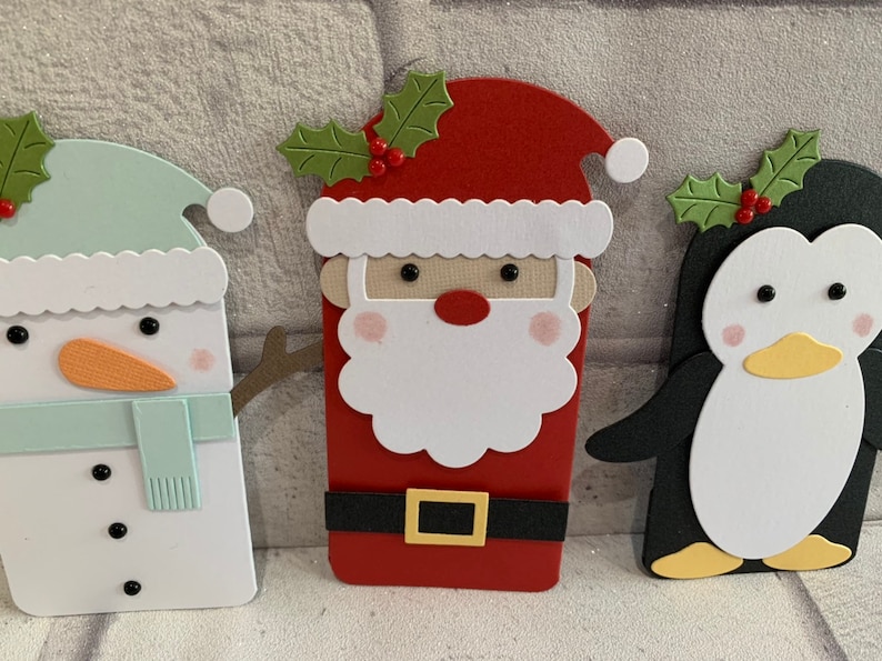Puede incluir: Tres recortes de papel con tem&aacute;tica navide&ntilde;a: un mu&ntilde;eco de nieve con gorro y bufanda verde menta, Pap&aacute; Noel con barba blanca y traje rojo, y un ping&uuml;ino con pico amarillo. Cada recorte tiene un dise&ntilde;o &uacute;nico y detalles festivos.