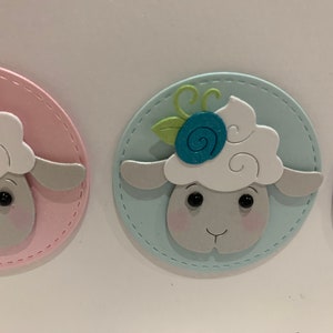 Op de afbeelding: Close-up van drie pastelkleurige schapenkopuitsparingen. Elk schaap heeft een pluizige witte kop, een grijs gezicht en zwarte ogen. De schapen zijn geplaatst op een roze, blauwe en paarse ovale achtergrond met een gestikte rand. E&eacute;n schaap heeft een blauwe bloem.