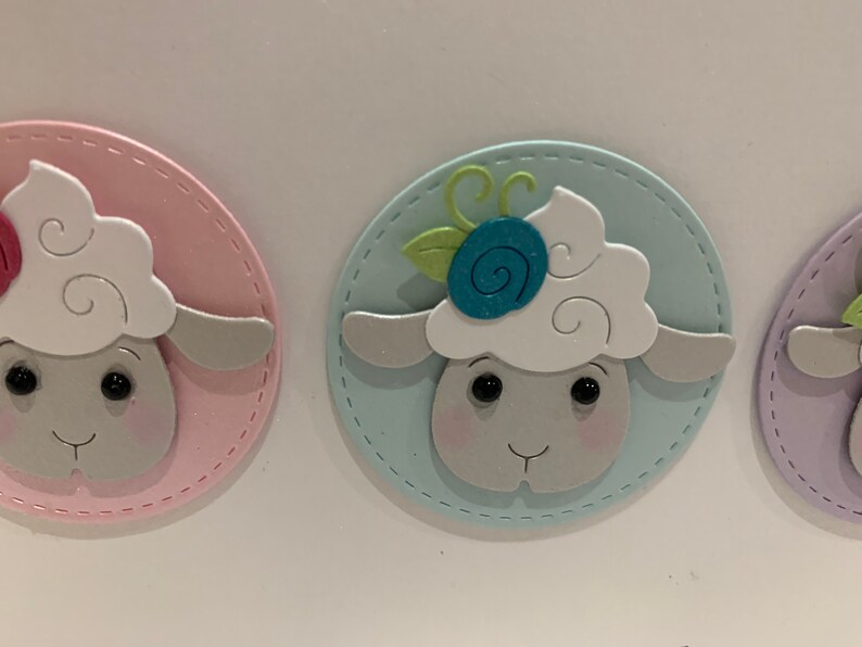 Op de afbeelding: Close-up van drie pastelkleurige schapenkopuitsparingen. Elk schaap heeft een pluizige witte kop, een grijs gezicht en zwarte ogen. De schapen zijn geplaatst op een roze, blauwe en paarse ovale achtergrond met een gestikte rand. E&eacute;n schaap heeft een blauwe bloem.