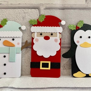 Puede incluir: Tres recortes de papel con tem&aacute;tica navide&ntilde;a: un mu&ntilde;eco de nieve con nariz de zanahoria y bufanda, Pap&aacute; Noel con barba blanca y cintur&oacute;n negro, y un ping&uuml;ino. Cada uno tiene una ramita de acebo encima.