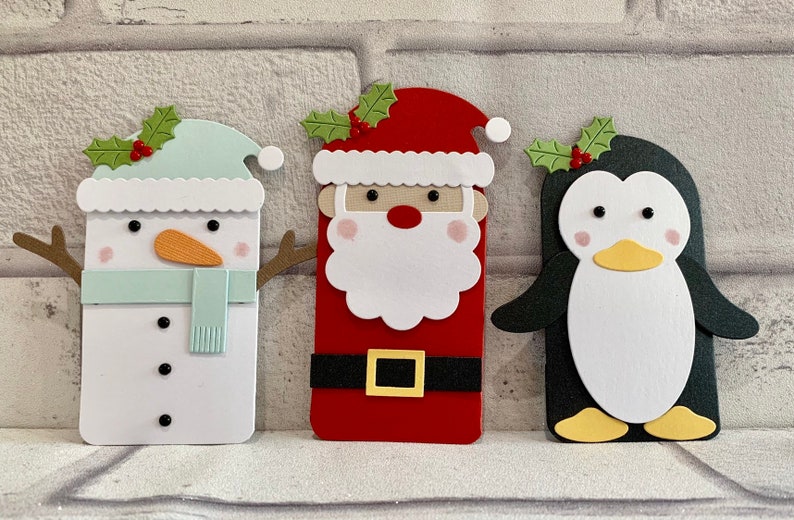 Puede incluir: Tres recortes de papel con tem&aacute;tica navide&ntilde;a: un mu&ntilde;eco de nieve con nariz de zanahoria y bufanda, Pap&aacute; Noel con barba blanca y cintur&oacute;n negro, y un ping&uuml;ino. Cada uno tiene una ramita de acebo encima.