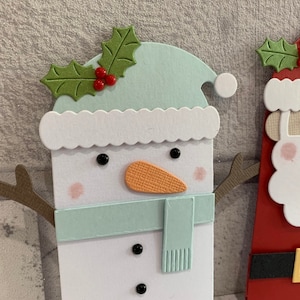 Puede incluir: Manualidad navide&ntilde;a con un mu&ntilde;eco de nieve y Pap&aacute; Noel. El mu&ntilde;eco de nieve tiene un sombrero y bufanda azul claro, una nariz naranja y ojos de bot&oacute;n negros. Pap&aacute; Noel tiene un traje rojo, un cintur&oacute;n negro con hebilla dorada y barba blanca. Ambos tienen adornos de acebo.