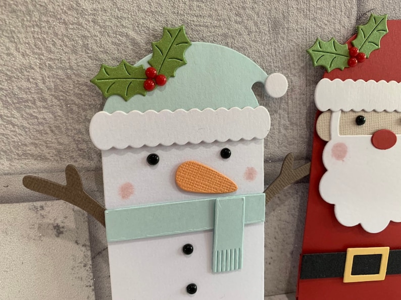 Puede incluir: Manualidad navide&ntilde;a con un mu&ntilde;eco de nieve y Pap&aacute; Noel. El mu&ntilde;eco de nieve tiene un sombrero y bufanda azul claro, una nariz naranja y ojos de bot&oacute;n negros. Pap&aacute; Noel tiene un traje rojo, un cintur&oacute;n negro con hebilla dorada y barba blanca. Ambos tienen adornos de acebo.