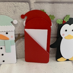 Puede incluir: Tres recortes de papel con tem&aacute;tica navide&ntilde;a: un mu&ntilde;eco de nieve, Pap&aacute; Noel y un ping&uuml;ino. El mu&ntilde;eco de nieve tiene un sombrero y una bufanda azul claro, Pap&aacute; Noel es rojo y el ping&uuml;ino es blanco y negro. Cada uno tiene detalles de acebo.