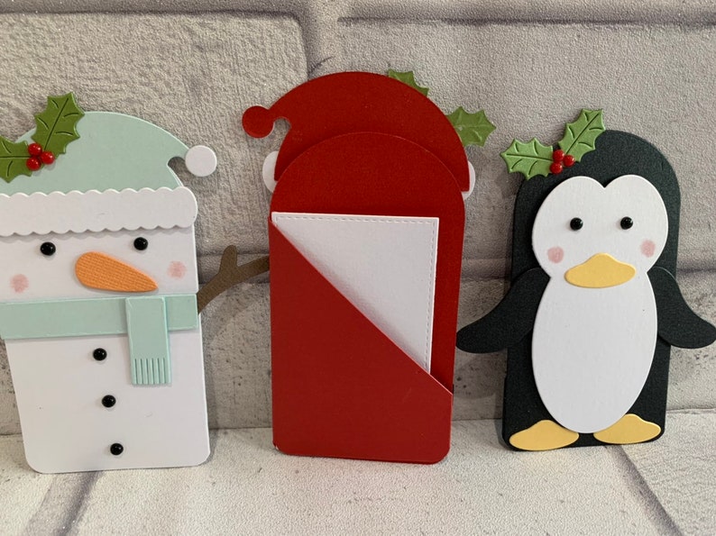 Puede incluir: Tres recortes de papel con tem&aacute;tica navide&ntilde;a: un mu&ntilde;eco de nieve, Pap&aacute; Noel y un ping&uuml;ino. El mu&ntilde;eco de nieve tiene un sombrero y una bufanda azul claro, Pap&aacute; Noel es rojo y el ping&uuml;ino es blanco y negro. Cada uno tiene detalles de acebo.