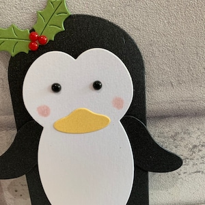 Puede incluir: Primer plano de un ping&uuml;ino de papel con cara blanca, cuerpo negro y pico amarillo. El ping&uuml;ino tiene mejillas rosadas y ojos negros. Una hoja de acebo verde con bayas rojas est&aacute; encima de su cabeza.