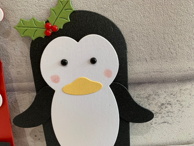 Puede incluir: Primer plano de un ping&uuml;ino de papel con cara blanca, cuerpo negro y pico amarillo. El ping&uuml;ino tiene mejillas rosadas y ojos negros. Una hoja de acebo verde con bayas rojas est&aacute; encima de su cabeza.
