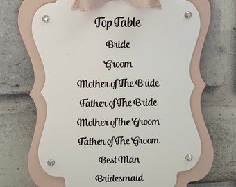 Puede incluir: Un letrero de mesa de boda con un diseño en capas rosa claro y blanco. El letrero presenta un lazo y adornos de cristal. El texto incluye "Top Table", "Bride", "Groom" y otros roles de la fiesta de bodas.