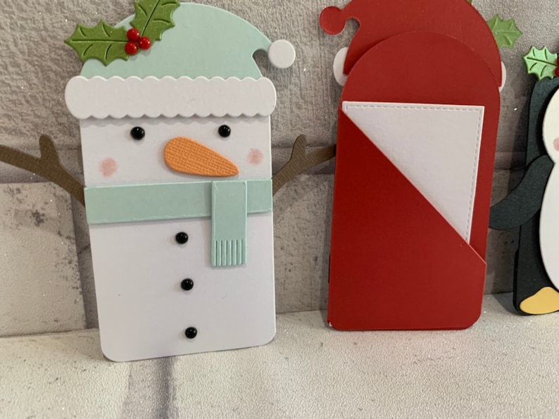 Puede incluir: Una colecci&oacute;n de dise&ntilde;os de tarjetas navide&ntilde;as hechas a mano. Las tarjetas presentan un mu&ntilde;eco de nieve con un gorro y bufanda verde menta, una tarjeta de Pap&aacute; Noel y una tarjeta de ping&uuml;ino. El mu&ntilde;eco de nieve tiene una nariz de zanahoria naranja y ojos de bot&oacute;n negros.