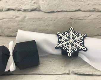 May include: A navy blue napkin ring in the shape of a snowflake with a white design and a small gem in the center. A white napkin is held by the ring. A navy blue cracker with a white ribbon is on the left.