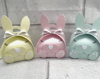 Könnte beinhalten: Drei pastellfarbene Geschenkboxen in Hasenform in Gelb, Rosa und Mintgrün. Jede Box hat eine weiße Schleife, lange Ohren und eine herzförmige Nase. Die Boxen haben einen Griff und sind mit Wimpern und Pfotenabdrücken verziert.