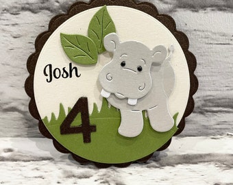Puede incluir: Manualidad de papel en capas con un hipopótamo gris, hojas verdes y el número 4. El número 4 es marrón. El nombre "Josh" está escrito en negro. El fondo es un círculo festoneado.