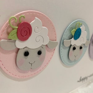 Op de afbeelding: Een close-up van drie pastelkleurige paasschapenhoofden. Elk schaap zit in een gestikt ovaal, de eerste in roze, de tweede in blauw en de derde in paars. De schapen hebben witte, donzige wol, roze wangen en bloemaccenten.