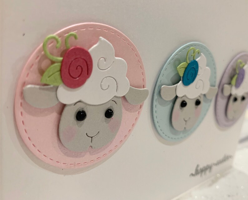Op de afbeelding: Een close-up van drie pastelkleurige paasschapenhoofden. Elk schaap zit in een gestikt ovaal, de eerste in roze, de tweede in blauw en de derde in paars. De schapen hebben witte, donzige wol, roze wangen en bloemaccenten.