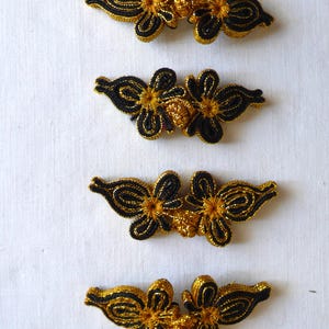 Pode incluir: Quatro fechos de rã florais pretos e dourados dispostos verticalmente sobre um fundo branco. Os fechos apresentam um design floral com fio preto delineando as pétalas e fio dourado preenchendo o centro. Estes fechos são provavelmente para roupas ou acessórios.