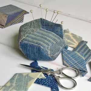 May include: A blue and white patchwork fabric pincushion with pearl-headed pins, surrounded by scraps of fabric.