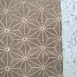 May include: Close-up of a brown fabric featuring a repeating geometric starburst pattern in off-white. The fabric has a textured appearance and a raw edge. The design is reminiscent of traditional Japanese motifs.