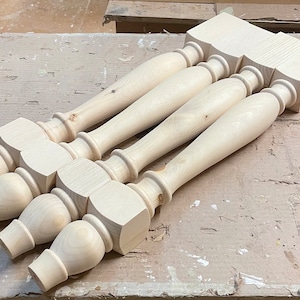 May include: Four unfinished wooden table legs with a turned design. The legs are stacked on top of each other and have a square base.