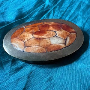 May include: An oval belt buckle featuring a polished brass frame surrounding a central inlay of segmented, multi-toned brown and tan pieces arranged in a geometric mosaic pattern that resembles a turtle shell design.