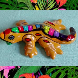 May include: A hand-painted ceramic lizard figurine in shades of yellow, orange, and green. The lizard has a colourful spine with rainbow-coloured segments. The figurine is decorated with white lines and red accents, and is a decorative item.