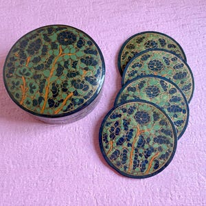 May include: A round, dark blue lidded box and four matching circular coasters. Each piece features a detailed floral pattern in shades of green, black, and orange, with thin orange stems and dark blue flower outlines on a muted green background.