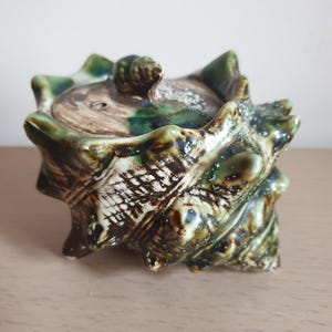 May include: A ceramic trinket box in the shape of a seashell. The box is glazed in green, brown, and white, with a textured surface. The lid has a small, rounded knob. The design is reminiscent of marine life.
