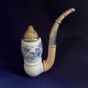 May include: A decorative pipe featuring a white ceramic bowl with a blue and white illustration of figures on horseback. The pipe has a gold-toned metal lid with a textured dome top and a curved, ribbed gold-toned metal stem attached to a cream-coloured base.