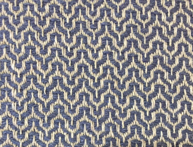 May include: A close-up of a woven fabric with a blue and beige geometric pattern. The fabric has a textured surface and a subtle sheen.