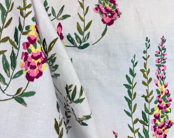 May include: White fabric with embroidered floral pattern. The design features pink, yellow, and green flowers and leaves. The fabric is folded, showing the texture and detail of the embroidery.