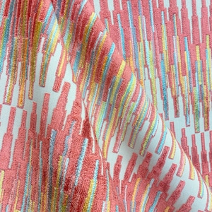 Op de afbeelding: Een close-up van een stof met een witte achtergrond en een patroon van verticale, kleurrijke strepen. De strepen zijn in tinten roze, blauw, geel en groen.