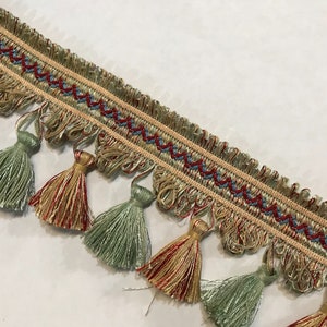 May include: A close-up of a decorative trim with a fringe of tassels in shades of green, red, and gold. The trim has a woven design with a repeating pattern of zigzags and lines.