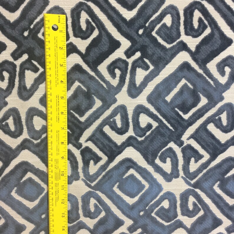 May include: Fabric with a repeating geometric pattern in shades of blue and gray. The design features stylized, swirling shapes within a square grid. A yellow ruler is placed vertically along the left side, showing measurements in inches.