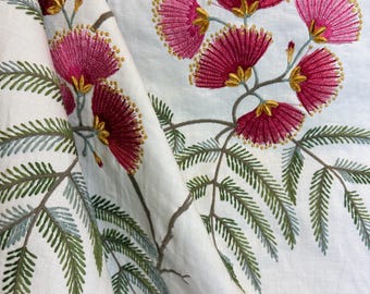 May include: White fabric with embroidered floral design. The embroidery features vibrant fuchsia flowers with gold accents and green fern-like leaves. The fabric is folded, showcasing the detailed craftsmanship and color contrast.