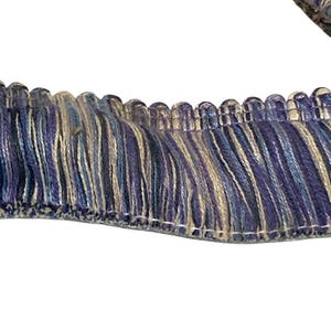 May include: Close-up of decorative fringe trim in shades of blue, cream, and gray. The trim features a scalloped edge and a textured, layered design, suitable for upholstery or craft projects.
