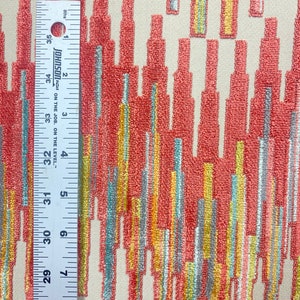 Op de afbeelding: Een close-up van een stof met een geometrisch patroon. Het patroon bestaat uit verticale strepen in tinten van koraal, geel, turquoise en grijs. De stof heeft een textuur.