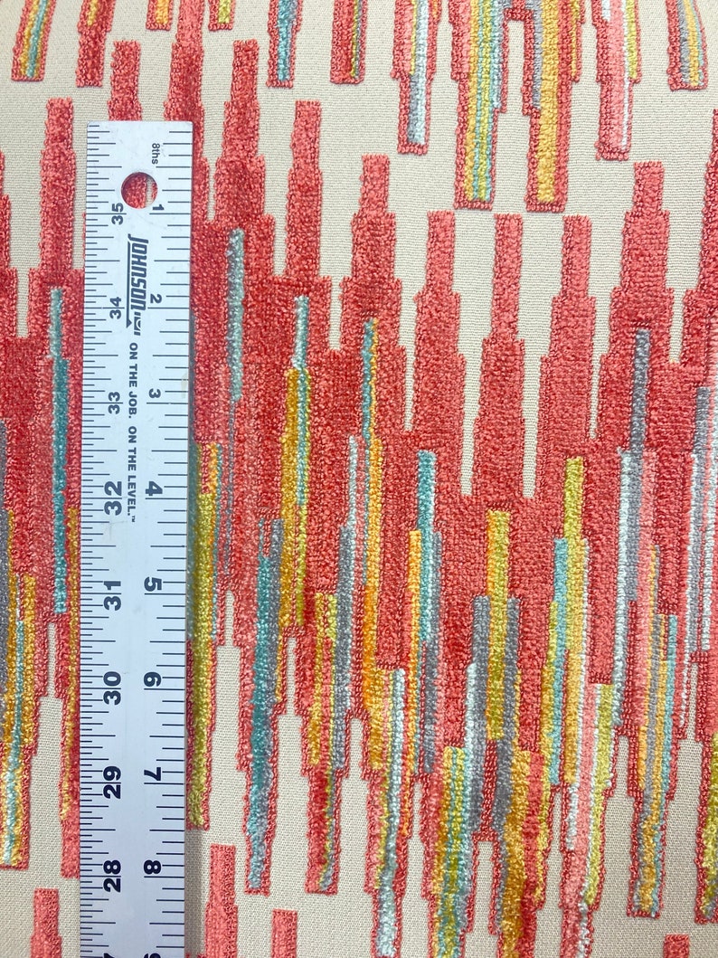 Op de afbeelding: Een close-up van een stof met een geometrisch patroon. Het patroon bestaat uit verticale strepen in tinten van koraal, geel, turquoise en grijs. De stof heeft een textuur.