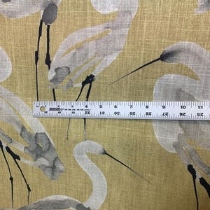 Pode incluir: Tecido amarelo-dourado com um padr&atilde;o repetido de gar&ccedil;as brancas e cinzentas. As gar&ccedil;as s&atilde;o retratadas em estilo aquarela, com pernas e bicos pretos. Uma r&eacute;gua &eacute; usada para medir o tecido.