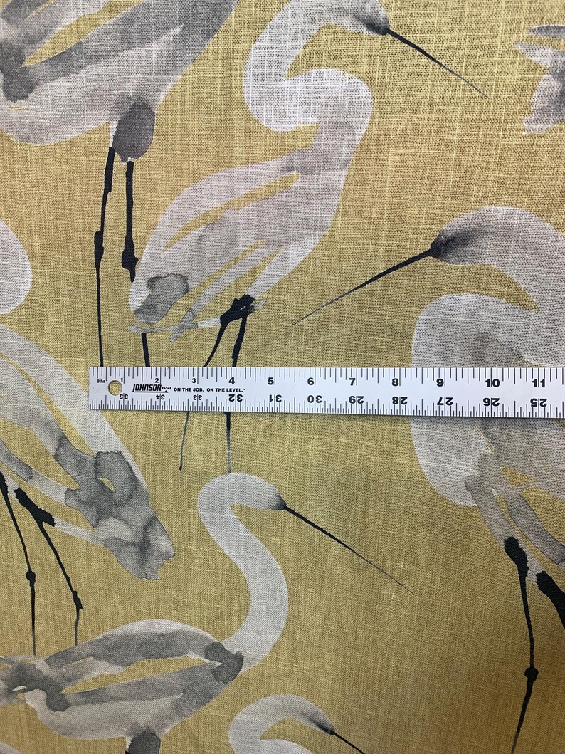 Pode incluir: Tecido amarelo-dourado com um padr&atilde;o repetido de gar&ccedil;as brancas e cinzentas. As gar&ccedil;as s&atilde;o retratadas em estilo aquarela, com pernas e bicos pretos. Uma r&eacute;gua &eacute; usada para medir o tecido.