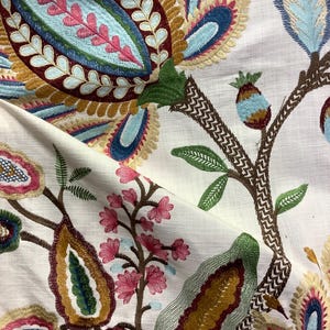May include: Close-up of white fabric with colorful embroidered floral design. The embroidery features large, stylized flowers in shades of blue, pink, green, and gold. The design includes detailed leaves and branches.