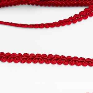 May include: A vibrant red gimp trim featuring a braided, scalloped edge design. The trim is displayed in multiple horizontal rows, showcasing the intricate woven texture and consistent deep red color throughout the entire length of the cord.